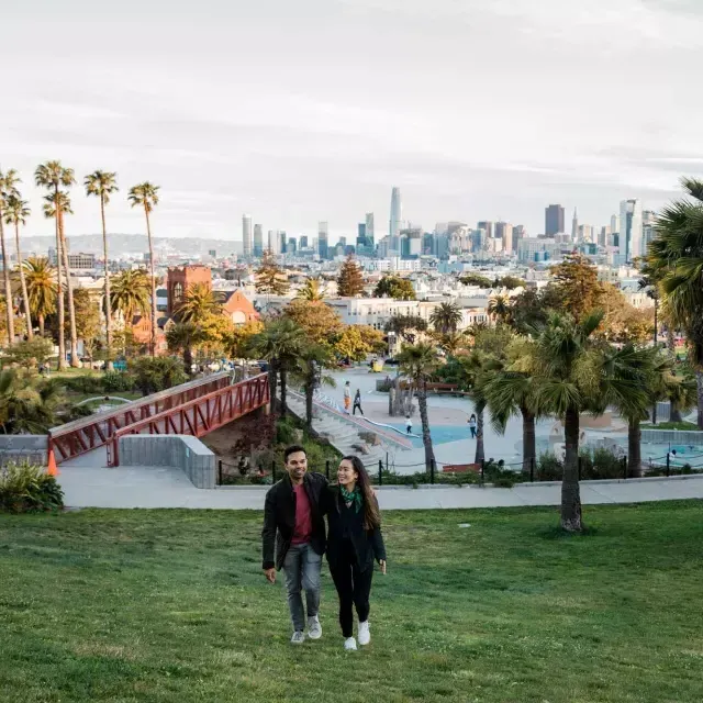 Una coppia cammina verso la telecamera, con Dolores Park e lo skyline di San Francisco alle spalle.